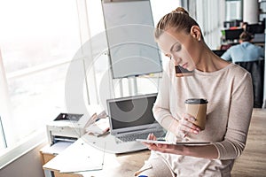 Beautiful girl is sitting at the adge of the table and looking at the tablet. Also she is talking on the phone and photo