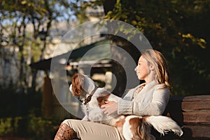 Beautiful girl is sitting on the bench in the park and enjoying with her dog Cavalier King Charles Spaniel