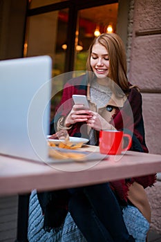 Beautiful girl sitting in cafe, using mobile phone and smiling happily. Outside. In the autumn.