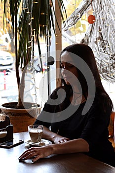 Beautiful girl is sitting in a cafe at a wooden table, enjoying drinking coffee from a glass cup. Silhouette in backlight from the