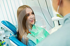 A beautiful girl is sitting in the dental chair and looking at a man doctor who is talking to her