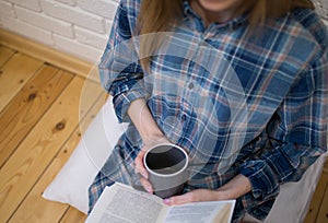 Beautiful girl is sitting on the floor in loft minimalist interior. Young woman in men`s checkered shirt is drinking coffee and