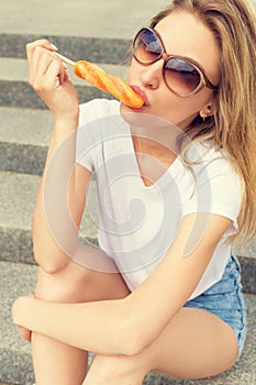 Beautiful girl sitting on the stairs in the town in sunglasses and denim shorts licking colorful ice lolly