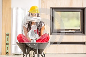 Beautiful girl sitting in a wheelbarrow inside the construction site with wearing overall and a hard hat, hiding face behind a