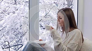 Beautiful girl on the sitting windowsill, drinking coffee and looking the window. Winter outside.