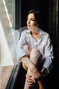 Beautiful girl sitting on the windowsill and sadly looking out the window