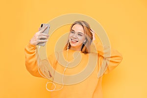 Beautiful girl with a smile makes a sephi and listens to music in the headphones on a yellow background, wearing a bright sweater