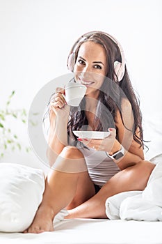 Beautiful girl smiles as she listen to the music via her headphones, drinking a cup of tea or coffee in the bed