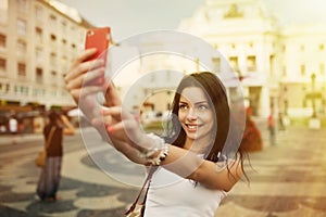 Krásne dievča s úsmevom a selfie. Mladá pekná žena sa baví a fotí sa s červeným smartfónom pred námestím