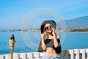 A beautiful girl speaks on the phone against the backdrop of the beach and ocean, sea. Woman in a big black hat and glasses on