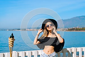 A beautiful girl speaks on the phone against the backdrop of the beach and ocean, sea. Woman in a big black hat and glasses on