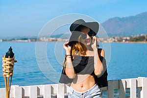 A beautiful girl speaks on the phone against the backdrop of the beach and ocean, sea. Woman in a big black hat and glasses on