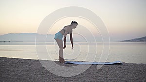 A beautiful girl spreads a karemat by the sea and begins to train. Sports, yoga, beach.