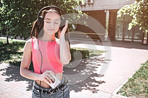 Beautiful girl is standing outside and listening to music in headphones. She is holding small and white player in right