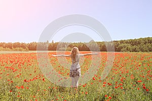 Beautiful girl is standing on summer field full of red poppy flowers in the grass. Sunny day with green lawn background. Happy