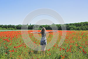 Beautiful girl is standing on summer field full of red poppy flowers in the grass. Sunny day with green lawn background. Happy