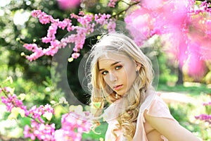 Beautiful girl standing under gentle tree with tiny hot pink blossom. Elegant young lady with long, blond, curled hair