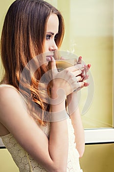 Beautiful girl standing at the window with a hot Cup of invigorating coffee early in the morning