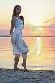 Beautiful girl stands on the beach, holds a red flower and looks into camera.