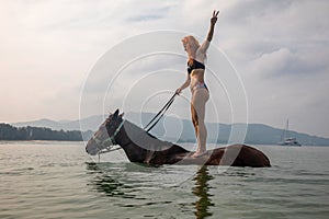 A beautiful girl stands on a horse in the water. Brown horse.