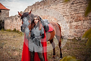 A beautiful girl stands next to a horse against the background of a medieval fortress. Fantasy costume, chain mail, cloak. A woman