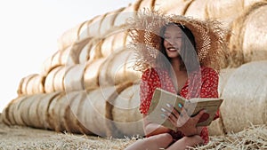 Beautiful girl in a straw hat is reading a book sitting in the hayloft. Education, literature and people concept.
