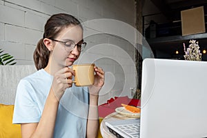 Beautiful girl student drinking coffee, tea, cocoa from a mug while sitting in front of a laptop. Business concept.