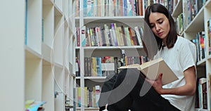 Beautiful girl student reading book, sitting among shelves with book on library floor, side view.