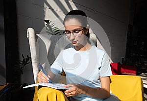 Beautiful girl student writes with a pen an assignment in a notebook while sitting on an armchair in a cafe