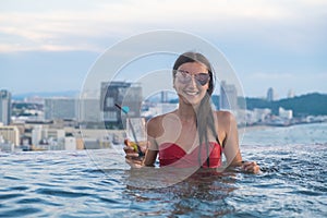 Beautiful girl in sunglasses with a cocktail in the pool with panoramic views of Pattaya city. Summer vacation holiday