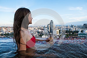 Beautiful girl in sunglasses with a cocktail in the pool with panoramic views of Pattaya city. Summer vacation holiday