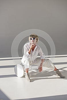 beautiful girl in sunglasses and fashionable white outfit sitting on chair and resting chin on hand