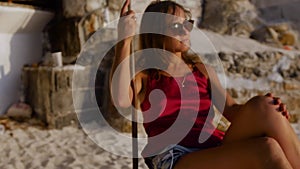 Beautiful girl at sunset swinging on a swing by the sea. Pretty young woman sitting on a swing and admiring the sunset