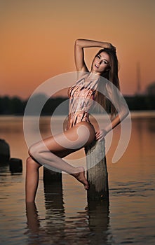 Beautiful girl in swimsuit posing provocatively during sunset at river shore. Attractive slim brunette woman sitting on pillar