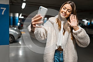 Beautiful girl takes selfie in the underground parking. Fashionable young woman with smartphone talking on video call