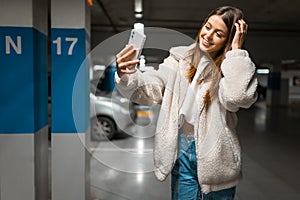 Beautiful girl takes selfie in the underground parking. Fasionable young woman with smartphone.