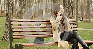 Beautiful girl talking on phone sitting on bench in autumn park smiling and enjoying conversation.