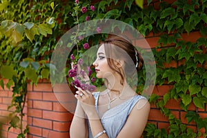 Beautiful girl in tender prom dress on brick wall and green ivy bush with flowers background. Female portrait