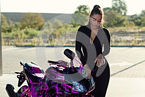 Beautiful girl in tight-fitting seductive suit washes black sport motorcycle and wipes it from magenta foam at sunrise.