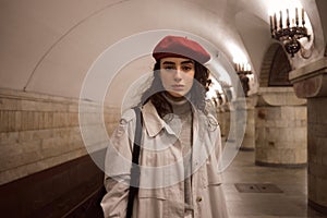 Beautiful girl in trench coat and red beret intently looking in camera waiting train at subway station