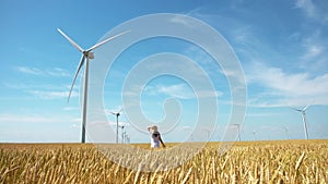 Beautiful girl walking on yellow field of wheat with windmills for electric power production