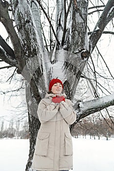Beautiful girl in warm clothes stands with her eyes closed under tree in winter park. Woman takes pleasure being in nature