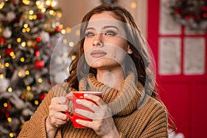 A beautiful girl in a warm sweater against the background of a Christmas tree and garlands is drinking coffee from a red mug.