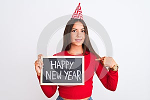 Beautiful girl wearing fanny party hat holding blackboard over isolated white background with surprise face pointing finger to