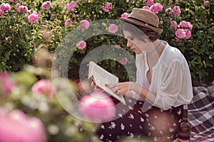 Beautiful girl wearing hat with book sitting on grass in rose gaden