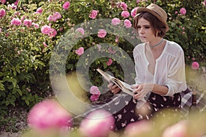 Beautiful girl wearing hat with book sitting on grass in rose gaden