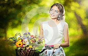 Beautiful girl wearing a nice white dress having fun in park with bicycle carrying a beautiful basket full of flowers. Vintage