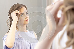 Beautiful girl with a wedding hairstyle looks at herself in the mirror, portrait of a young girl. beautiful make-up.