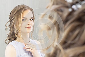 Beautiful girl with a wedding hairstyle looks at herself in the mirror, portrait of a young girl. beautiful make-up.