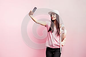 Beautiful girl in white cap and with a backpack on her shoulders dressed in pink t-shirt and black jeans is doing selfie